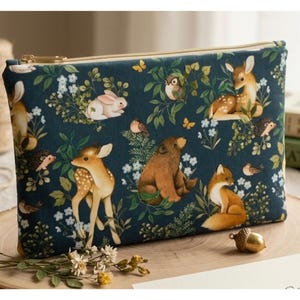 May include: A zippered pouch with a dark teal background featuring a woodland scene. The design includes illustrations of deer, rabbits, foxes, birds, and a bear amidst floral and foliage elements. The pouch has a gold zipper.