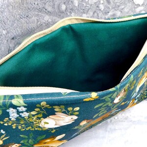 May include: A teal zippered pouch with a woodland animal print featuring rabbits, hedgehogs, and foliage. The interior is a solid emerald green. The zipper is beige.