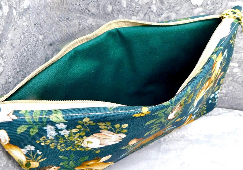 May include: A teal zippered pouch with a woodland animal print featuring rabbits, hedgehogs, and foliage. The interior is a solid emerald green. The zipper is beige.