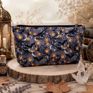 May include: A zippered pouch featuring a dark gray fabric pattern with black silhouettes of bare tree branches, flying bats, white crescent moons, small stars, and scattered orange autumn leaves. The pouch has a black zipper closure at the top.