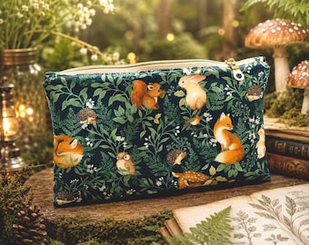 Può includere: Una pochette rettangolare con cerniera in tessuto verde scuro, decorata con un motivo boschivo di volpi, scoiattoli, conigli, ricci e gufi immersi tra foglie verdi, felci e piccoli fiori bianchi, rifinita con una zip in metallo dorato.
