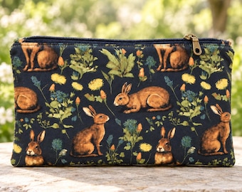 Può includere: Una pochette rettangolare con chiusura a zip su base in tessuto blu scuro, caratterizzata da un motivo ripetuto di conigli marroni, fiori di tarassaco gialli e fogliame verde. La pochette presenta un cursore metallico in alto a destra.