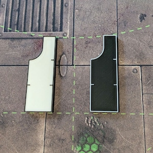 Op de afbeelding: Twee spelbordstukken, één wit en één zwart, met een unieke vorm. Elk stuk heeft een rechthoekige basis en een gebogen bovenkant, met zwarte lijnen voor meting. De stukken staan tegen een bruine en grijze achtergrond.