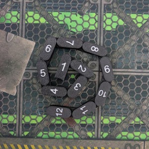 Può includere: Una collezione di pezzi di gioco neri, oblunghi, con numeri bianchi da 1 a 12 disposti in cerchio. I pezzi sono su una superficie strutturata, verde e grigia, con un motivo geometrico.