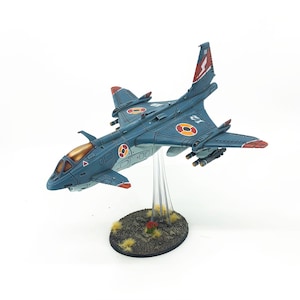 Può includere: Modello dettagliato di un jet da combattimento blu e rosso. Il modello presenta dettagli intricati, tra cui segni circolari arancioni e bianchi, un fulmine rosso sulla coda e piccoli attacchi missilistici. Il jet è montato su un supporto trasparente.