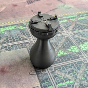 Puede incluir: Un objeto negro impreso en 3D con una base bulbosa y una parte superior circular con cuatro pestañas salientes. El objeto tiene una superficie texturizada y parece ser un componente o parte de un mecanismo más grande. El artículo está sobre una superficie estampada.