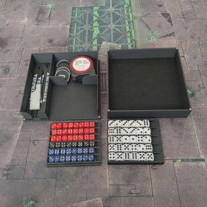 Puede incluir: Un organizador de juegos gris con múltiples compartimentos. Contiene dados rojos, blancos y azules, fichas circulares y una cinta métrica. El organizador está abierto, revelando su contenido, y se encuentra sobre una superficie estampada.