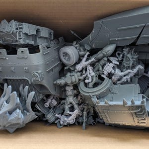 May include: A box containing unassembled grey plastic model kit components. The parts include vehicle sections, figures, and other detailed pieces, indicating a miniature wargame or hobby project.
