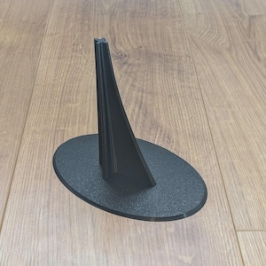 May include: A black display stand with an oval base and a tall, angled support. The stand is made of a textured material and is designed to hold an item. The stand is on a wooden surface.