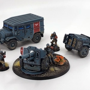 Puede incluir: Modelos militares en miniatura, que incluyen un camión azul, un cañón, un remolque y varios soldados. Los modelos están pintados en tonos azules y rojos, con detalles y texturas. La escena se desarrolla sobre una superficie blanca.