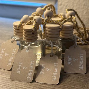 May include: A group of small, clear glass bottles with light-colored wooden caps and hanging cords featuring gold metallic threads. Each bottle has a light brown paper tag attached with the white handwritten text LEATHER & LACE on the front.