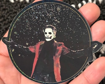 May include: A circular sticker featuring a figure in a black outfit with a red jacket, white face paint, and outstretched arms against a starry background. The sticker has a holographic border.