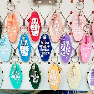 May include: A selection of vibrant keyrings, each with a unique design and phrase. Keyrings feature text such as "Self Love Club" and "Drive Safe Bestie." Each keyring has a tassel and is attached to a metal ring.
