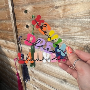 May include: A rainbow-coloured acrylic bookmark with the words "Dream Big" written in black. The bookmark has a tassel on each side, one pink and one blue.