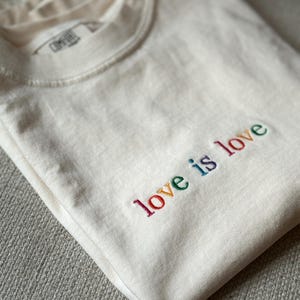 May include: Cream-coloured t-shirt with the embroidered phrase "love is love" in rainbow colours. The shirt is neatly folded, displaying the text clearly. The fabric appears soft and comfortable.