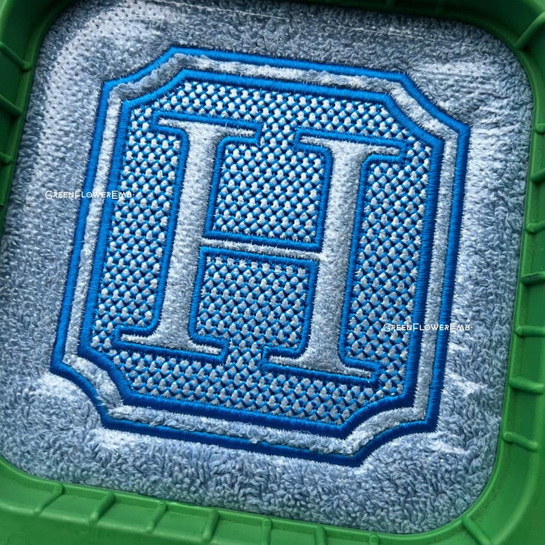 May include: A close-up of a blue and silver embroidered letter "H" on a light blue fabric. The letter is surrounded by a decorative border.