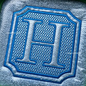 May include: A square embroidered patch featuring a large, silver-colored letter H centered within a blue-bordered frame. The background of the letter is filled with a textured, light blue cross-stitch pattern, all set against a fuzzy gray fabric.