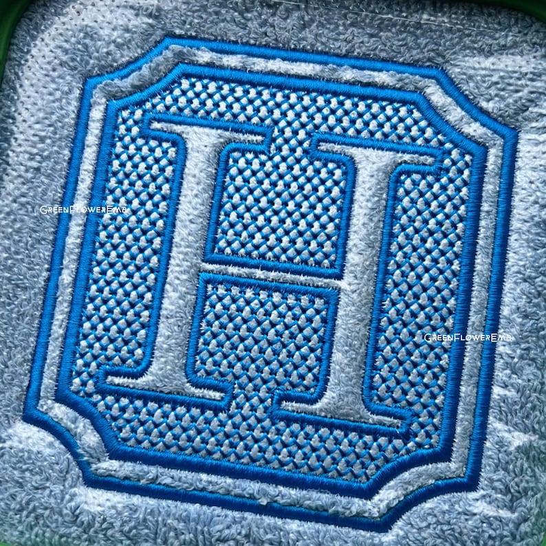 May include: A square embroidered patch featuring a large, silver-colored letter H centered within a blue-bordered frame. The background of the letter is filled with a textured, light blue cross-stitch pattern, all set against a fuzzy gray fabric.