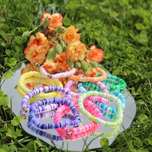 Puede incluir: Una colección de pulseras de cuentas coloridas en un arcoíris de colores, incluyendo amarillo, naranja, rosa, morado, azul y verde. Las pulseras están dispuestas sobre una superficie de espejo plateado, con un ramo de flores naranjas en el fondo.
