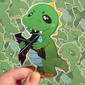 May include: A green cartoon dinosaur sticker with a black lightning bolt in its mouth. The dinosaur has big eyes and a yellow crest on its head.