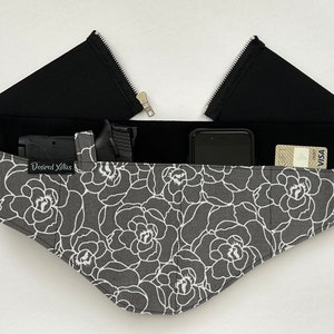 May include: A black fabric belt with a zipper closure and multiple pockets. The belt has a floral print with white flowers on a grey background. The text "Desired Xotic" is visible on the belt.