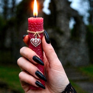Peut inclure: Une bougie rouge allumée avec une surface texturée, attachée avec de la ficelle et un charm en forme de cœur argenté. La bougie est tenue devant un arrière-plan extérieur flou. La flamme est jaune et orange vif.