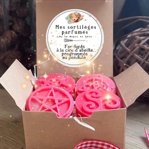 Peut inclure: Une boîte en carton contenant quatre fondants parfumés roses avec des paillettes. Les fondants présentent divers motifs, dont un pentagramme et des volutes. La boîte porte une étiquette circulaire avec le texte français : "Mes sortilèges parfumés".