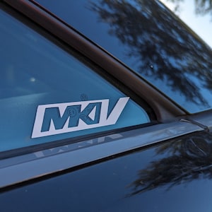 Op de afbeelding: Een witte sticker met de tekst "MKV" op een zwart autovenster. De sticker zit op de achterruit van de auto.