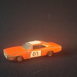 May include: Orange die-cast toy car with a white roof and the number "01" on the side. The vintage toy car has black wheels and is set against a dark background. The car's paint shows signs of wear.