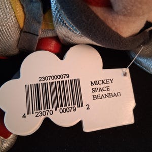 May include: A close-up of a Mickey Space Beanbag plush toy. The toy has a tag with a barcode and the words "MICKEY SPACE BEANBAG". The toy is primarily gray, red, and yellow.
