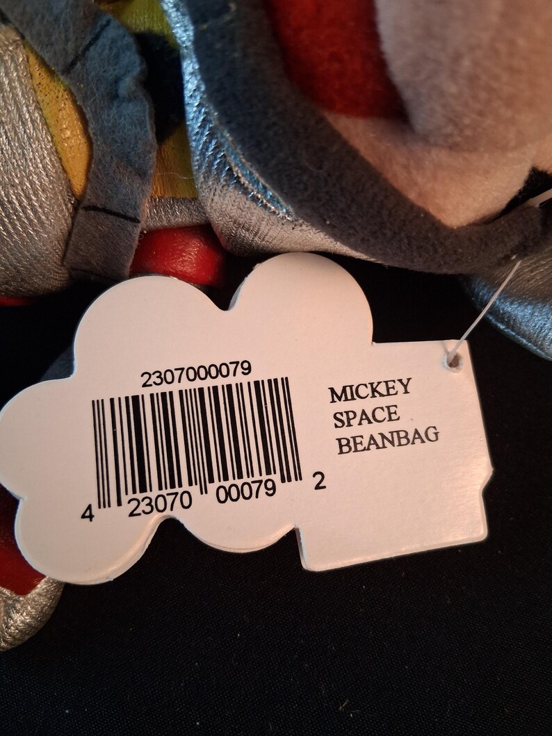 May include: A close-up of a Mickey Space Beanbag plush toy. The toy has a tag with a barcode and the words "MICKEY SPACE BEANBAG". The toy is primarily gray, red, and yellow.