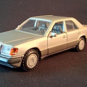 May include: A silver Mercedes-Benz model car with a two-tone paint scheme. The car has a detailed front grille, headlights, and wheels. The interior features blue seats. The model is set against a dark background.