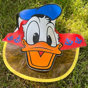 May include: A yellow plastic sun visor with a cartoon image of Donald Duck wearing a blue sailor hat and red and white striped shirt. The visor has a clear plastic shield and anchors printed on the sides.