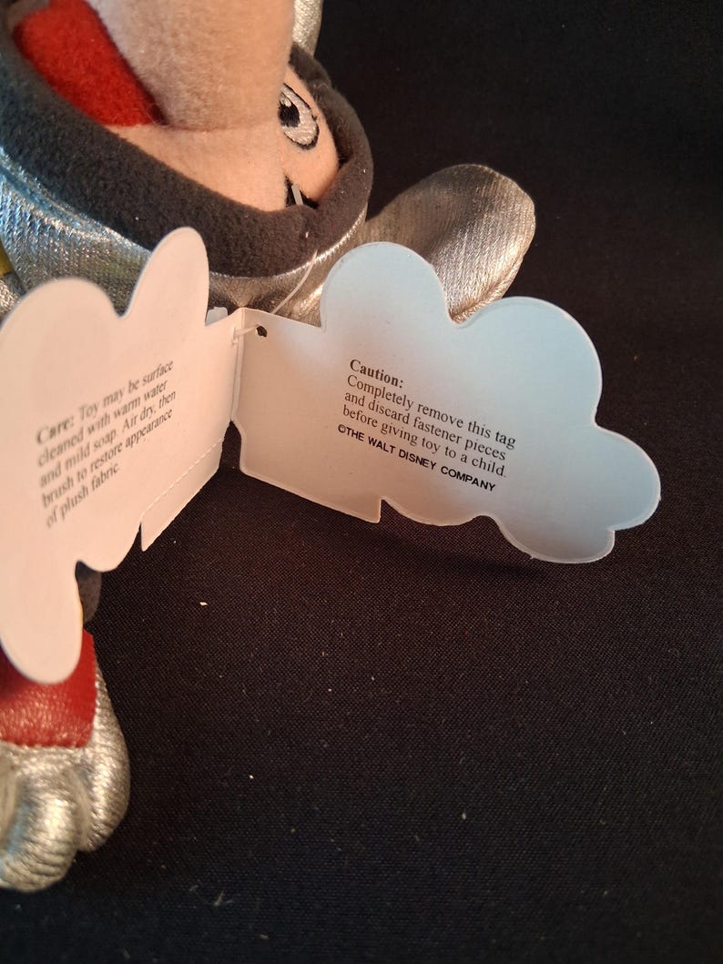 May include: A plush toy with a silver and black design, featuring a red mouth and a tag with care instructions and a caution notice from The Walt Disney Company.