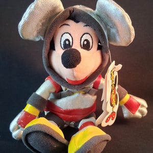 May include: A plush Mickey Mouse toy wearing a silver hooded outfit with red and yellow accents. The toy has a black nose, big eyes, and a red smile. A tag is attached to the toy.