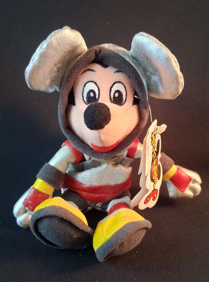 May include: A plush Mickey Mouse toy wearing a silver hooded outfit with red and yellow accents. The toy has a black nose, big eyes, and a red smile. A tag is attached to the toy.
