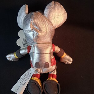 May include: A plush toy mouse in a silver astronaut suit with red and yellow accents. The toy has large ears and a tag attached. The mouse is approximately 6 inches tall.