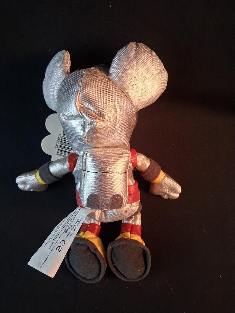 May include: A plush toy mouse in a silver astronaut suit with red and yellow accents. The toy has large ears and a tag attached. The mouse is approximately 6 inches tall.