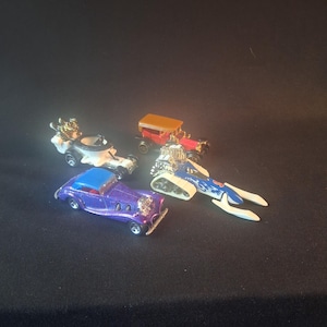 Può includere: Una collezione di modellini di auto in vari colori, tra cui viola, rosso e blu. Le auto hanno design unici, alcune con dettagli cromati e caratteristiche personalizzate. Le modellini di auto sono disposte su una superficie scura.