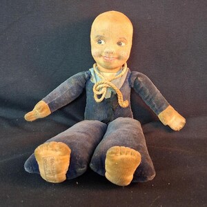 May include: A vintage cloth doll with a bald head, painted facial features, and a smiling expression. The doll wears a dark blue velvet suit with a light blue collar and a decorative rope tie around the neck. Tan fabric hands and feet are visible.
