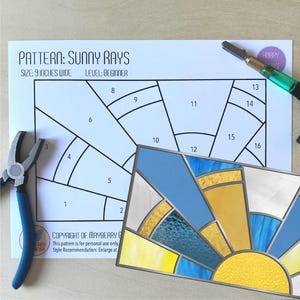 May include: A stained glass pattern for a sunny rays design. The pattern is printed on white paper and includes numbered pieces for a 9-inch wide project. The finished stained glass piece is shown below the pattern.