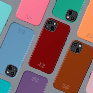 May include: A collection of colorful phone cases in various shades, including blue, pink, and orange. Each case features a unique set of initials engraved on the back. The cases are arranged on a gray surface, showcasing their glossy finish and modern design.