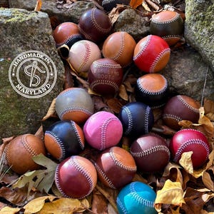 Op de afbeelding: Een verzameling ronde leren honkballen in diverse kleuren zoals rood, blauw, roze, beige, bruin en zwart, elk met witte stiksels. Eén bal is voorzien van de tekst "BABBLING BOOTS" en een rond logo met de tekst "HANDMADE IN MARYLAND".