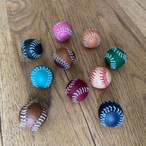 Puede incluir: Una colección de diez pequeñas pelotas esféricas estilo béisbol en varios colores, incluyendo rosa, bronceado, negro, marrón, verde, verde azulado, granate y crema. Cada pelota presenta un patrón de costura blanco distintivo en la superficie.