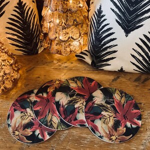 May include: Four round coasters with a floral design featuring red, orange, and green leaves on a black background. The coasters are on a wooden surface, with decorative pillows and a gold-colored tree in the background.