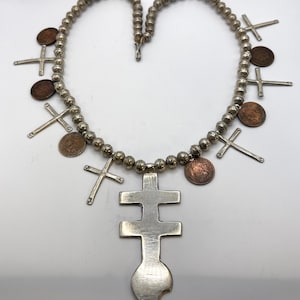May include: A silver necklace with a beaded chain, small crosses, and copper coins. The pendant is a large, double-barred cross with a flame-shaped base. The necklace is a unique piece of jewellery.