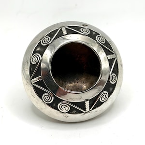 Puede incluir: Un recipiente esférico plateado con una gran abertura central. El recipiente está decorado con una banda de diseños geométricos, incluyendo espirales y formas angulares. El diseño está grabado en el metal, creando un patrón contrastante oscuro y claro.