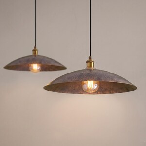 May include: Two hanging pendant lights with a rustic, metallic finish. Each light features a wide, shallow shade with a warm, glowing bulb. The fixtures have brass-colored accents and black cords, creating a vintage industrial aesthetic.