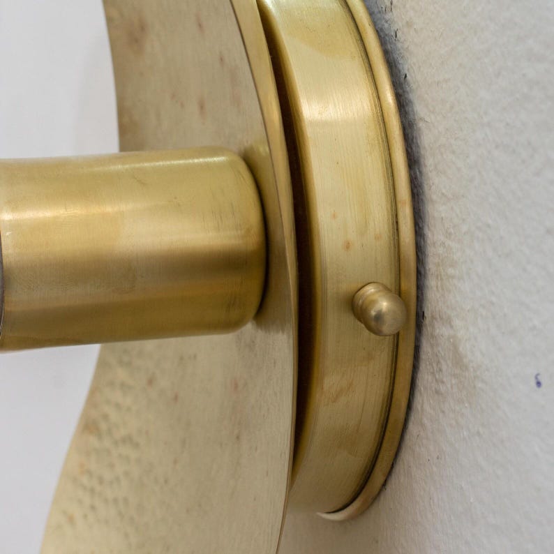 Puede incluir: Primer plano de un aplique de pared de lat&oacute;n. El aplique presenta una placa trasera circular y un brazo cil&iacute;ndrico, todo en un c&aacute;lido tono dorado. El dise&ntilde;o es simple y elegante, con una est&eacute;tica vintage.