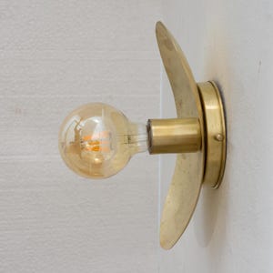 Puede incluir: Aplique de pared de lat&oacute;n con un dise&ntilde;o curvo decorativo. El aplique tiene una base redonda y un brazo cil&iacute;ndrico que sostiene una bombilla de estilo vintage. La bombilla emite un brillo &aacute;mbar c&aacute;lido, y el accesorio est&aacute; montado en una pared blanca.